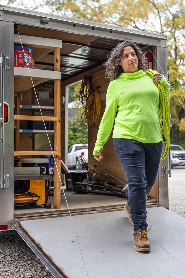 Woman exiting a mobile workshop trailer wearing Dovetail Workwear's Christa DIY Pant in Sulfur Navy with a bright neon yellow pullover and work boots