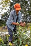 Woman tending to yellow flowers in a garden wearing the Dovetail Workwear Givens Work Shirt in Indigo Stripe, paired with an orange cap