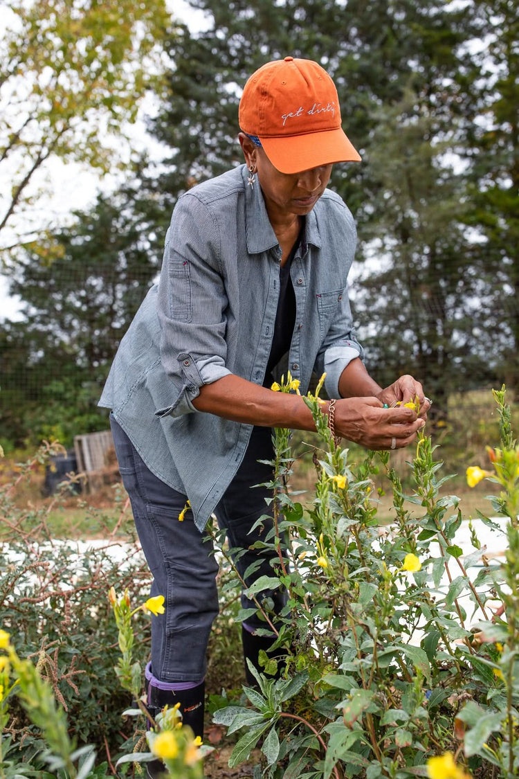 Woman tending to yellow flowers in a garden wearing the Dovetail Workwear Givens Work Shirt in Indigo Stripe, paired with an orange cap