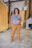 Anna Maternity Work Pants in Saddle Brown Canvas by Dovetail Workwear shown on a woman standing in a workshop wearing a gray "Work Like a Mother" tee and tan work pants with reinforced knees