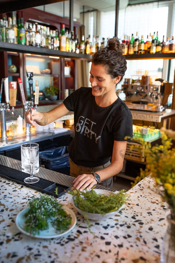 A woman wearing the black Get Dirty™ Crew Neck Tee by Dovetail Workwear behind a bar, pouring a drink with fresh herbs on the counter