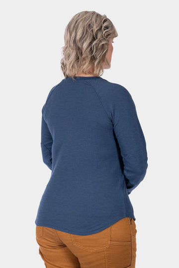 Back view of a woman wearing the Dovetail Workwear Honed Long Sleeve Thermal Henley in Atlantic Blue with Kodiak Brown pants, showcasing the reinforced elbows and waffle knit thermal base layer.