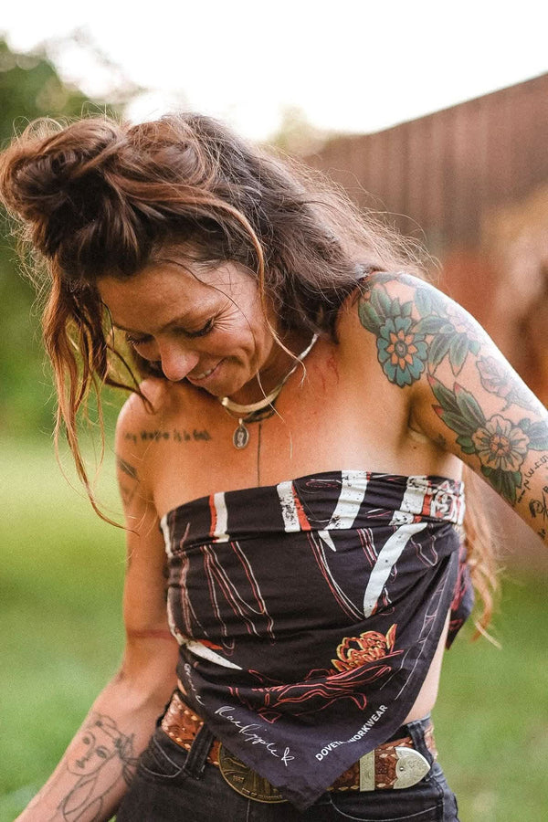 Rae Ripple x Dovetail Workwear Brave Wings Bandana in black tied around a woman's neck, displaying hand-drawn wing and floral artwork and text against an outdoor backdrop