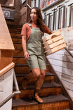 Woman wearing Dovetail Workwear Utility Short Overalls in Light Kelp Green descending wooden stairs while carrying wooden barrel bands, showcasing the shortall's relaxed fit and practical design