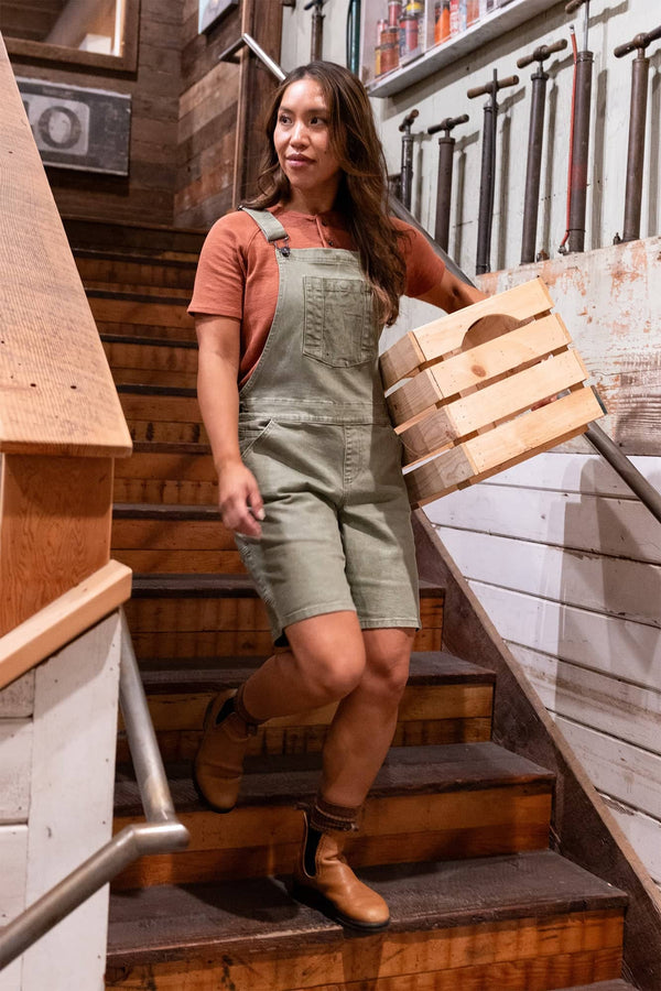 Woman wearing Dovetail Workwear Utility Short Overalls in Light Kelp Green descending wooden stairs while carrying wooden barrel bands, showcasing the shortall's relaxed fit and practical design
