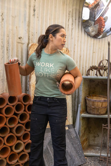 Woman wearing Dovetail Workwear GO TO "Work Like a Mother" Graphic Work Tee in sage green with black work pants, holding terracotta pipes in an industrial setting