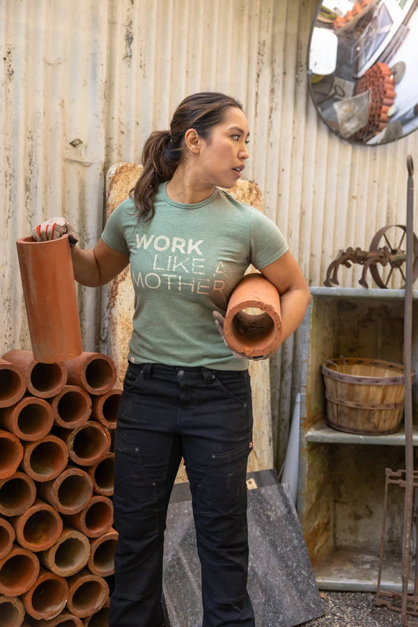 Woman wearing Dovetail Workwear GO TO "Work Like a Mother" Graphic Work Tee in sage green with black work pants, holding terracotta pipes in an industrial setting