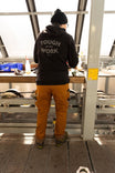 Back view of a woman wearing the Dovetail Workwear GO TO™ Graphic Hoodie in Carbon Black with tan work pants, standing at a workbench with large windows behind her