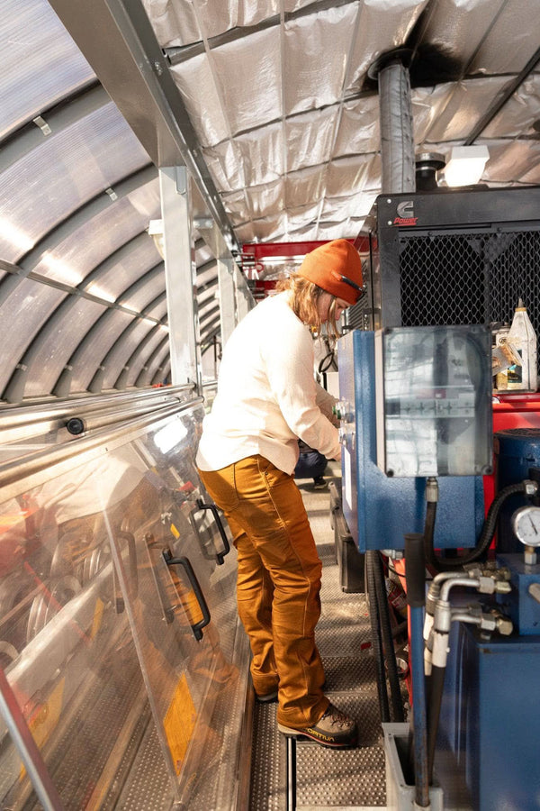 Woman wearing Dovetail Workwear's Britt Utility HOTSWAP™ Pant in Saddle Brown Thermal Denim, demonstrating the thermal denim's durability in an industrial work setting