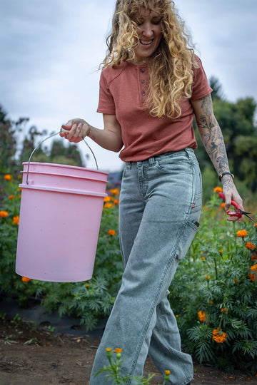Woman wearing Dovetail Workwear's Old School Summer Henley in Terracotta, harvesting flowers in a garden with a pink bucket and pruning shears