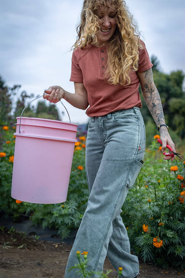 Woman wearing Dovetail Workwear's Old School Summer Henley in Terracotta, harvesting flowers in a garden with a pink bucket and pruning shears