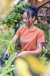 Woman wearing Dovetail Workwear's GO TO "Get Dirty" Graphic Work Tee in rust, tending to potted plants in a lush garden setting