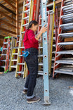 Woman inspecting the blue level on Dovetail Workwear Old School High Rise Graphene Denim Pant in Heather Black, demonstrating the high-rise fit and straight-leg silhouette among stored ladders