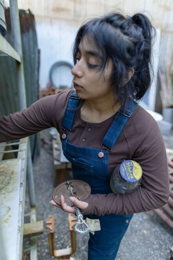 Woman wearing Dovetail Workwear's Honed Long Sleeve Thermal Henley in Kodiak Brown with waffle knit thermal base layer and reinforced elbows, styled with denim overalls in an outdoor work setting
