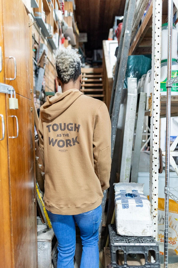 Back view of a woman wearing the Saddle Brown GO TO™ Graphic Hoodie by Dovetail Workwear, displaying the graphic on back, in a warehouse setting