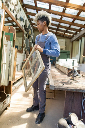 Woman holding a window frame in a workshop, wearing the Dovetail Workwear GO TO™ Crew Sweatshirt in Slate Blue/Atlantic Blue with brown apron over it
