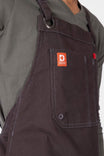 Close-up detail of the Dovetail Workwear Obana Work Apron in Kodiak Brown, showing the orange brand label, front pocket, and cross-back strap on a woman wearing the apron