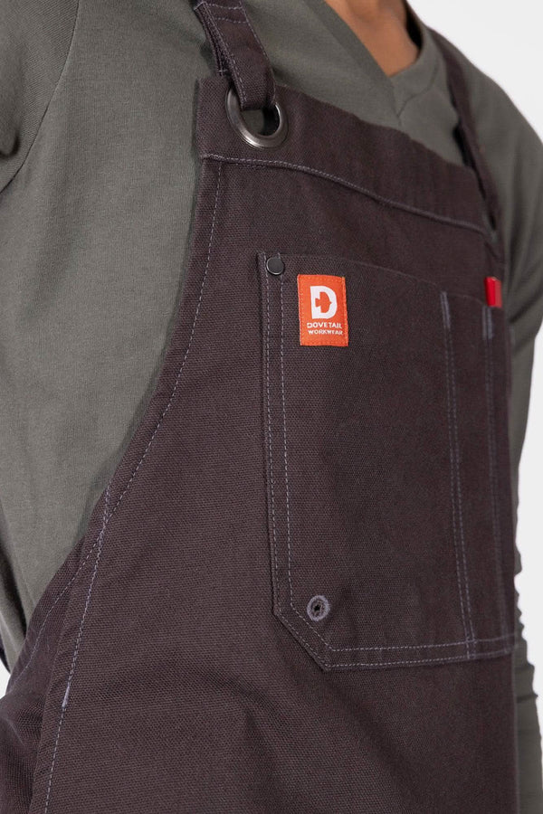 Close-up detail of the Dovetail Workwear Obana Work Apron in Kodiak Brown, showing the orange brand label, front pocket, and cross-back strap on a woman wearing the apron