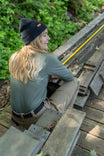 Black Dovetail Beanie worn by a woman sitting on wooden steps, shown from the side with long blonde hair and green foliage in background