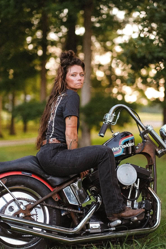 Back view of a woman sitting on a Harley-Davidson motorcycle wearing John Deere X Dovetail Workwear Into Town Bootcut Jeans in Classic Black