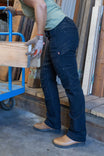 Woman wearing Dovetail Workwear's Britt Utility Pant in No Fade Black Canvas, demonstrating the nested pockets and straight leg fit while carrying wood on a blue dolly
