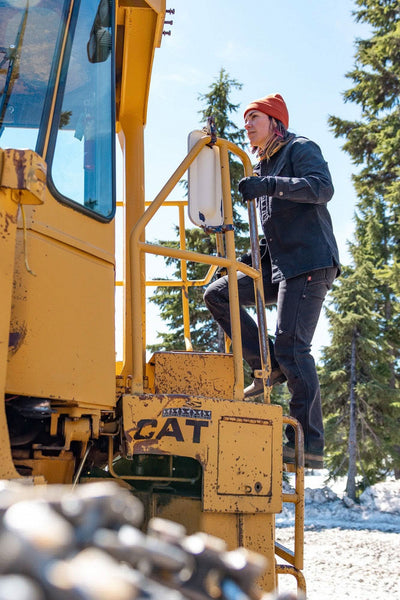 Woman wearing Dovetail Workwear Britt Utility HOTSWAP™ Pant in Black Thermal Denim climbing onto yellow CAT equipment in snowy conditions