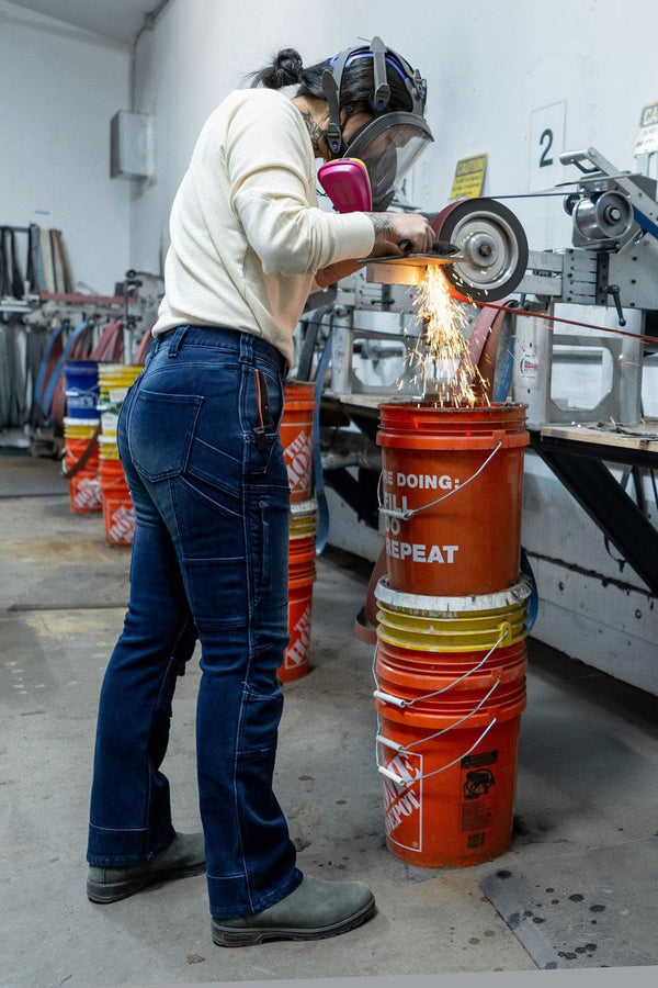 Woman wearing indigo Britt X Graphene Denim Pant by Dovetail Workwear while operating grinding equipment in a workshop, demonstrating durability in heavy-duty work conditions