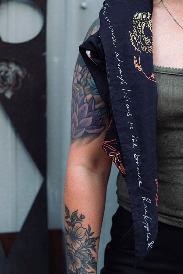 Rae Ripple x Dovetail Workwear Brave Wings Bandana in black styled as a wild rag around a woman's arm, displaying hand-drawn wing and flower artwork and inspirational text