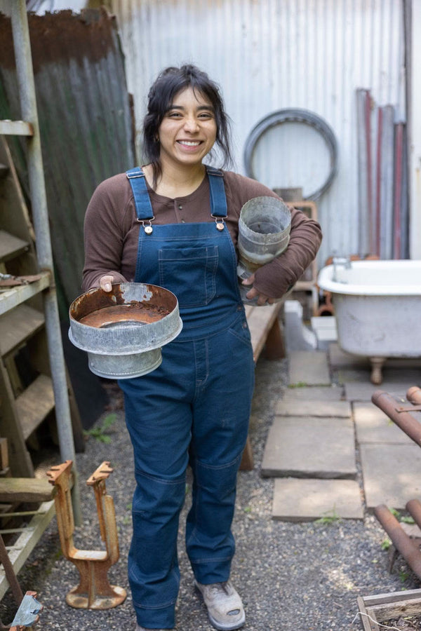 Woman wearing Dovetail Workwear GO TO™ Stretch Canvas Overalls in Atlantic Blue, holding galvanized metal buckets in a workshop setting