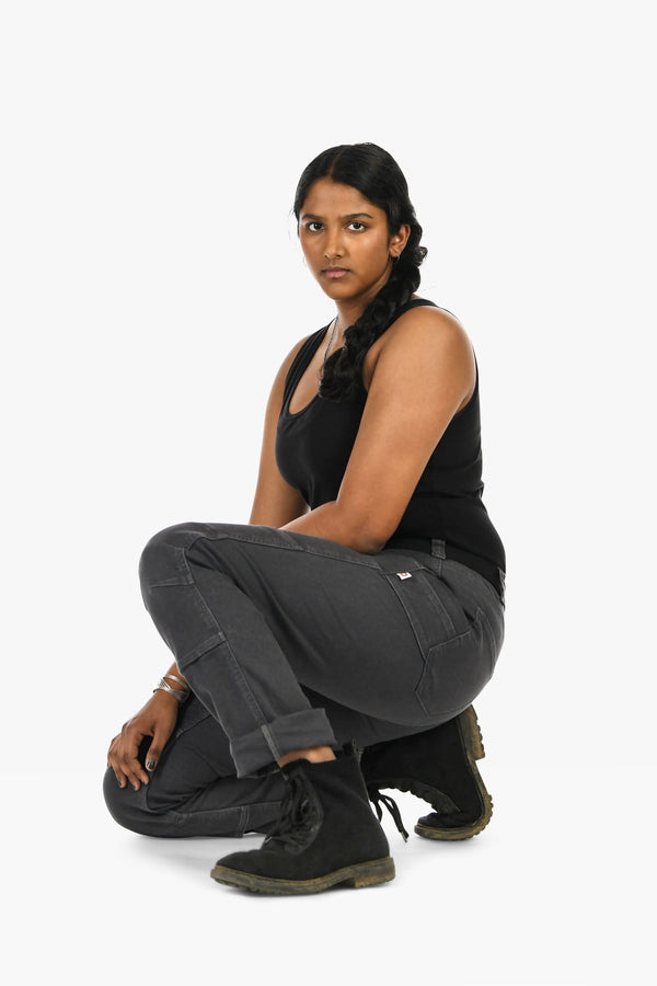 Woman crouching in Dovetail Workwear GO TO™ Stretch Canvas Pant in Cement Grey, showcasing the straight leg fit and tool loop detail from a side angle