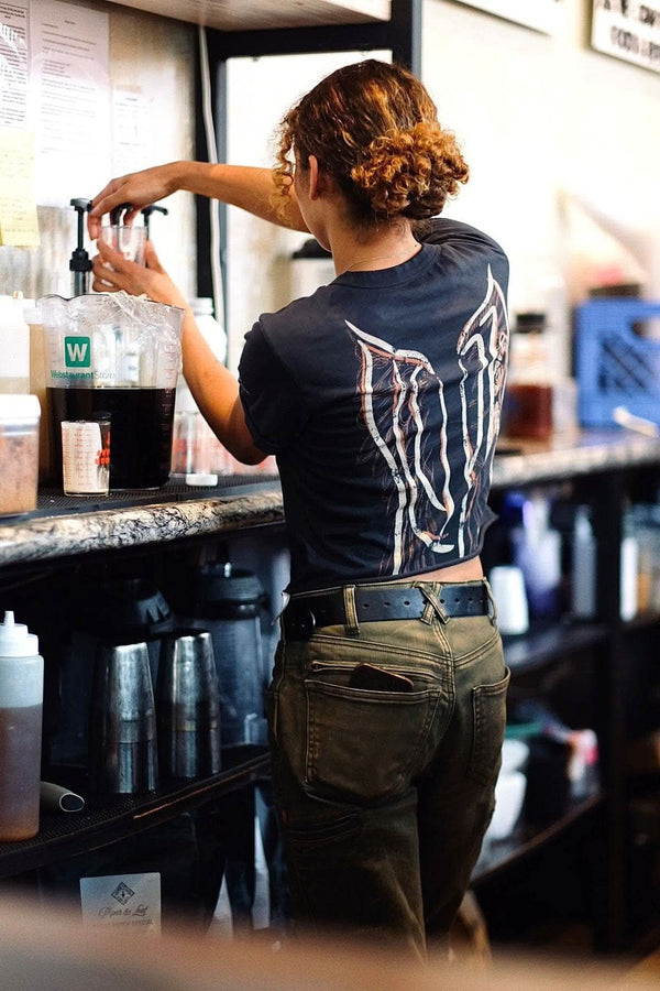 Back view of a woman wearing the Rae Ripple x Dovetail Workwear Brave Wings T-Shirt in black, featuring signature wings design across the back and relaxed fit 100% cotton construction.