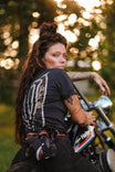 Back view of a woman wearing the Rae Ripple x Dovetail Workwear Brave Wings T-Shirt in black, featuring signature winged artwork and sitting on a motorcycle at golden hour