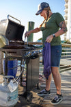 Woman wearing Dovetail Workwear Wabash Work Short in Wabash Stripe while grilling on a waterfront deck, showcasing the shorts' Hip Slot™ Tool loop and practical design for outdoor work
