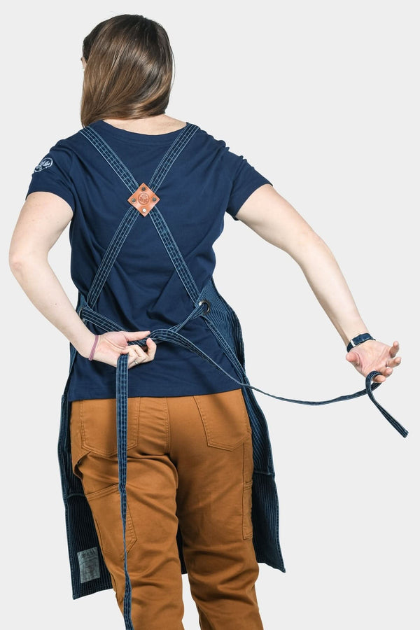 Back view of a woman wearing the Dovetail Workwear Obana Work Apron in Dark Blue Wabash Stripe, showing cross-back straps and tool pockets