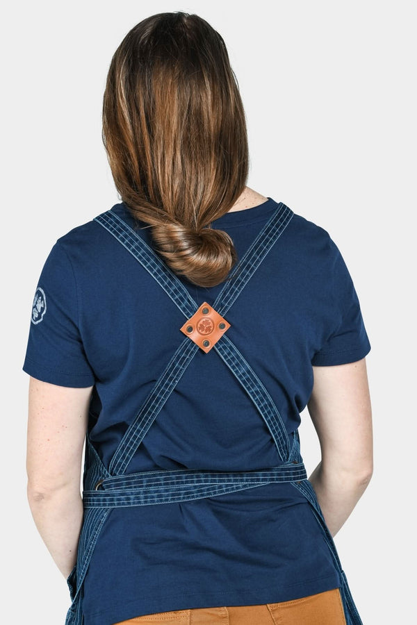 Back view of a woman wearing the Dovetail Workwear Obana Work Apron in Dark Blue Wabash Stripe, displaying cross-back straps and leather accents