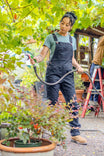 Woman watering plants in Dovetail Workwear's GO TO™ Stretch Canvas Overalls in Carbon Black, showcasing the durable work overalls' practical design and comfortable fit for outdoor tasks