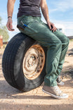 Woman wearing Aventurine Green Christa X DIY COOLSWAP™ Pant by Dovetail Workwear, sitting on a tire, showcasing the slim leg fit and COOLSWAP™ ripstop fabric durability