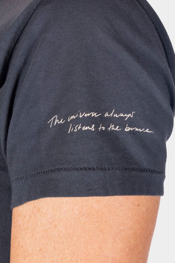 Close-up detail of the Rae Ripple x Dovetail Workwear Brave Wings T-Shirt in black, showing the handwritten mantra "The universe always listens to the brave" printed on the left sleeve