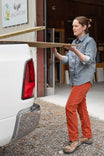 Woman wearing Dovetail Workwear's Anna Taskpant in Paprika Canvas, demonstrating the straight leg fit and Hip Slot™ Tool loop while holding lumber outside a workshop