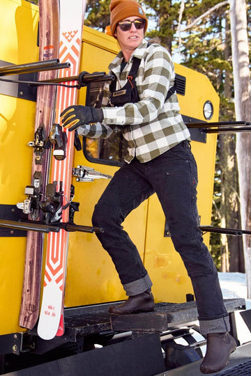 Woman wearing Dovetail Workwear's Britt Utility Pant in Black Stretch Thermal Denim with reinforced knees, standing on a yellow ski lift carrying skis in a snowy mountain setting