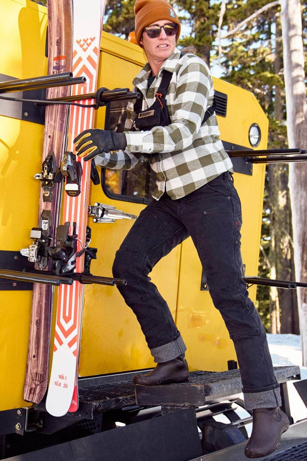 Woman wearing Dovetail Workwear's Britt Utility Pant in Black Stretch Thermal Denim with reinforced knees, standing on a yellow ski lift carrying skis in a snowy mountain setting