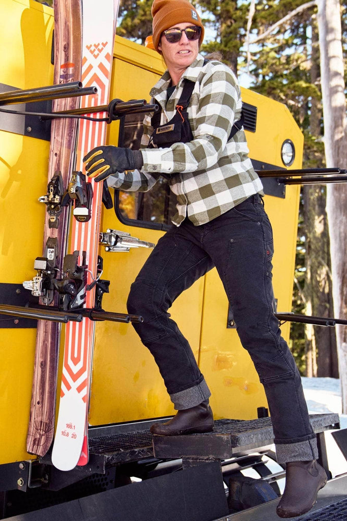 Woman wearing Dovetail Workwear's Britt Utility Pant in Black Stretch Thermal Denim with reinforced knees, standing on a yellow ski lift carrying skis in a snowy mountain setting
