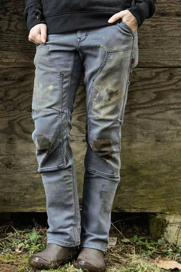 Back view of a woman wearing Dovetail Workwear's Britt Utility Pant in Grey Stretch Thermal Denim, showing reinforced knees and straight leg fit against a wooden backdrop