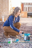Woman crouching while holding a paint can, wearing Dovetail Workwear's Britt Utility Pant in Saddle Brown Canvas with a blue jacket, showcasing tool loop and pocket functionality on a job site