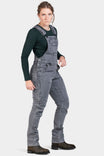 Woman wearing Dovetail Workwear's Freshley Drop Seat Overalls in grey stretch thermal denim with dark long-sleeve shirt and brown boots, shown from the side