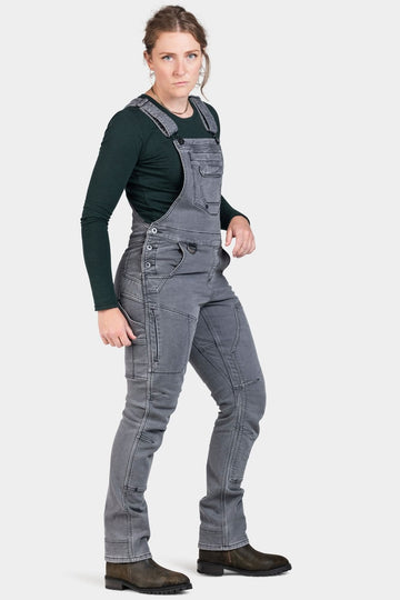 Woman wearing Dovetail Workwear's Freshley Drop Seat Overalls in grey stretch thermal denim with dark long-sleeve shirt and brown boots, shown from the side