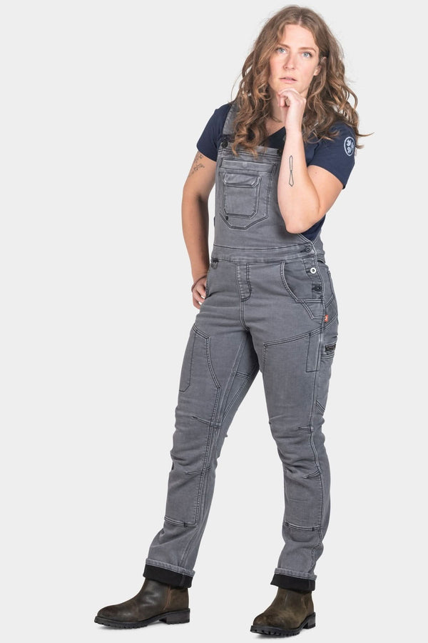 Woman wearing Dovetail Workwear Freshley Drop Seat Overalls in grey stretch thermal denim with reinforced knees and tool loops, paired with a navy shirt and dark boots