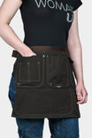 Dovetail Tool Apron in Kodiak Brown worn by a woman, showing multiple front pockets with contrast stitching, adjustable waist belt with quick release buckle, and dual hammer loops