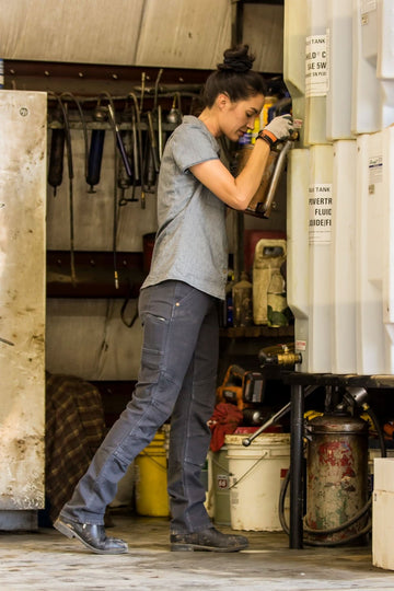 Woman wearing Dovetail Workwear GO TO™ Stretch Canvas Pant in Cement Grey working in an industrial setting, demonstrating the straight leg fit and tool loop functionality