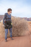 Woman wearing Dovetail Workwear Dovetail Tool Apron in Kodiak Brown with multiple pockets and hammer loops, standing outdoors in desert landscape